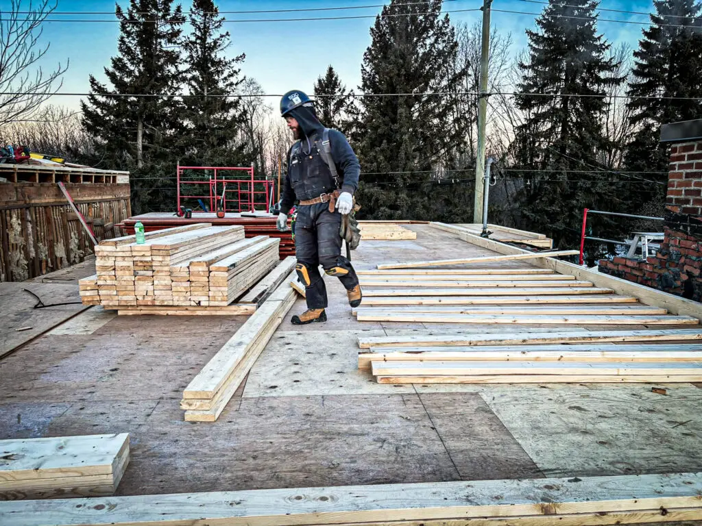 Construction worker in safety gear standing on wooden framing structure of a residential house addition under cloudy skies. He scans a pile of well-organized lumber for the perfect peice