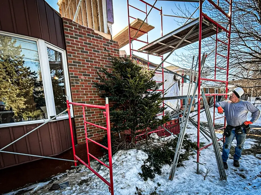 Construction worker in a hard hat and safety gear setting up scaffolding on a residential renovation site in winter.